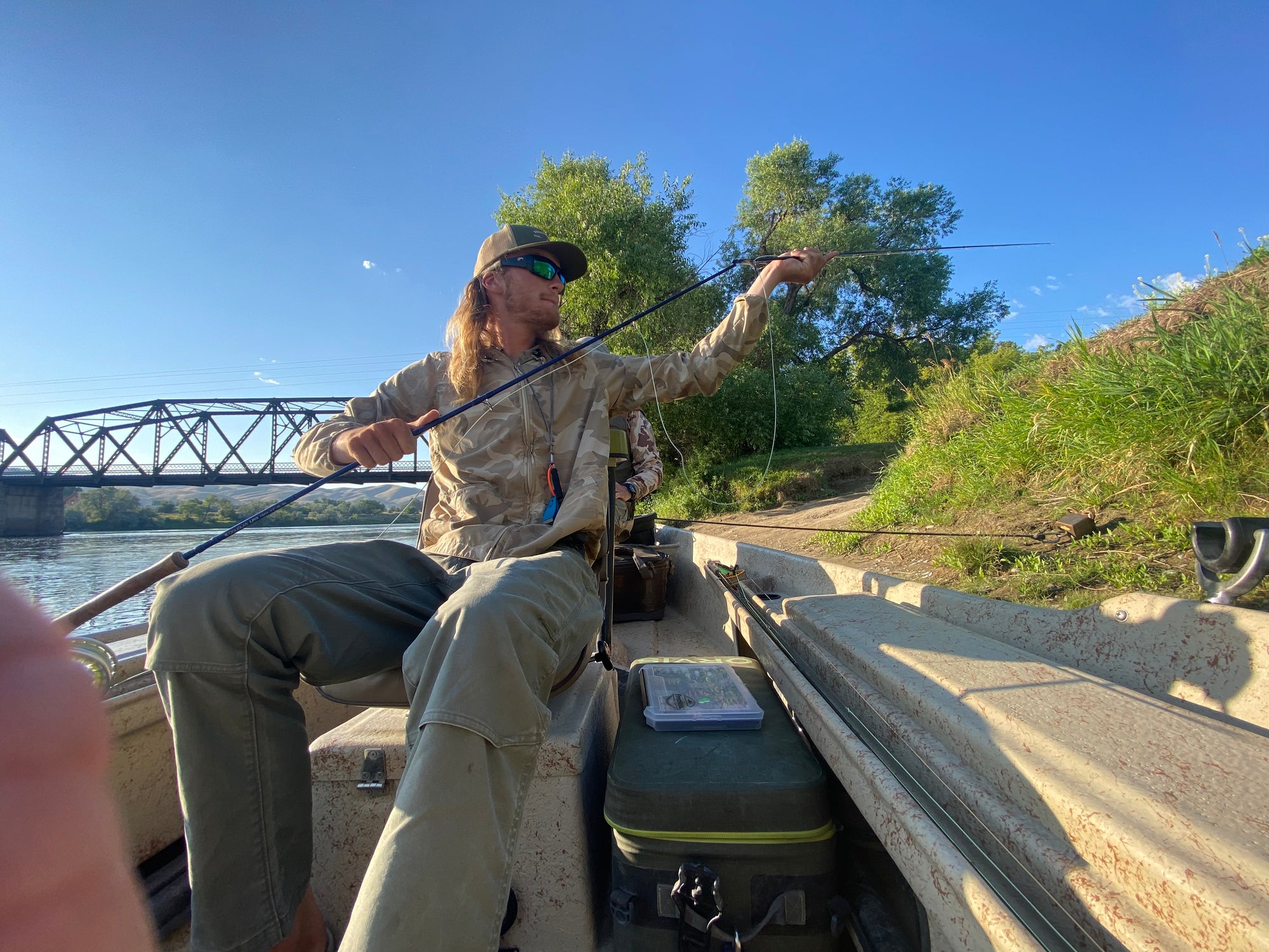 Man sitting in boat holding fly fishing rod near riverbank with green trees and blue sky behind