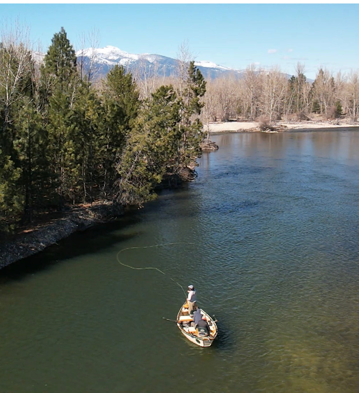 A lone angler casting a fly line from a drift boat on a river bordered by pine and leafless trees with snowy mountains beyond