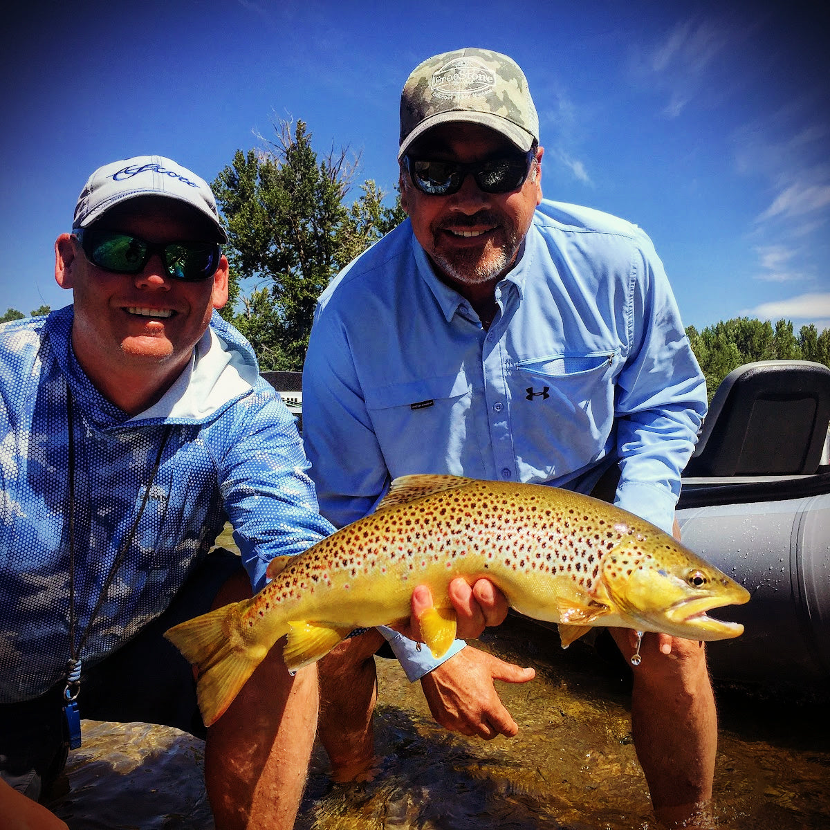 Two anglers smiling while holding a large spotted brown trout near an inflatable boat on a sunny day
