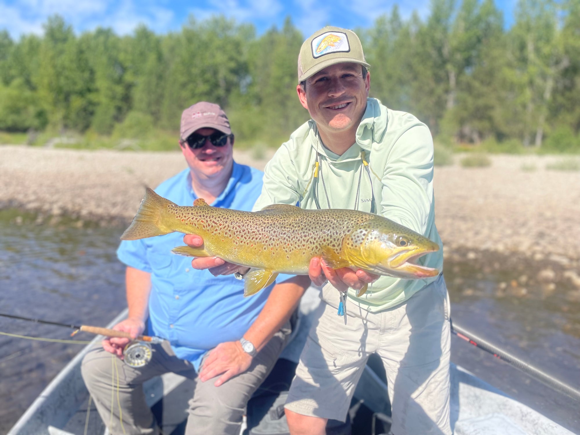Man in light shirt holding large brown trout while another seated angler holds fishing rod by riverbank trees