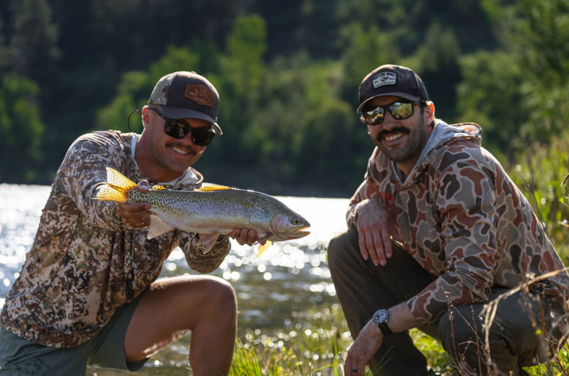 Two men wearing sunglasses and camo clothing posing near a river with a large trout on a sunny day