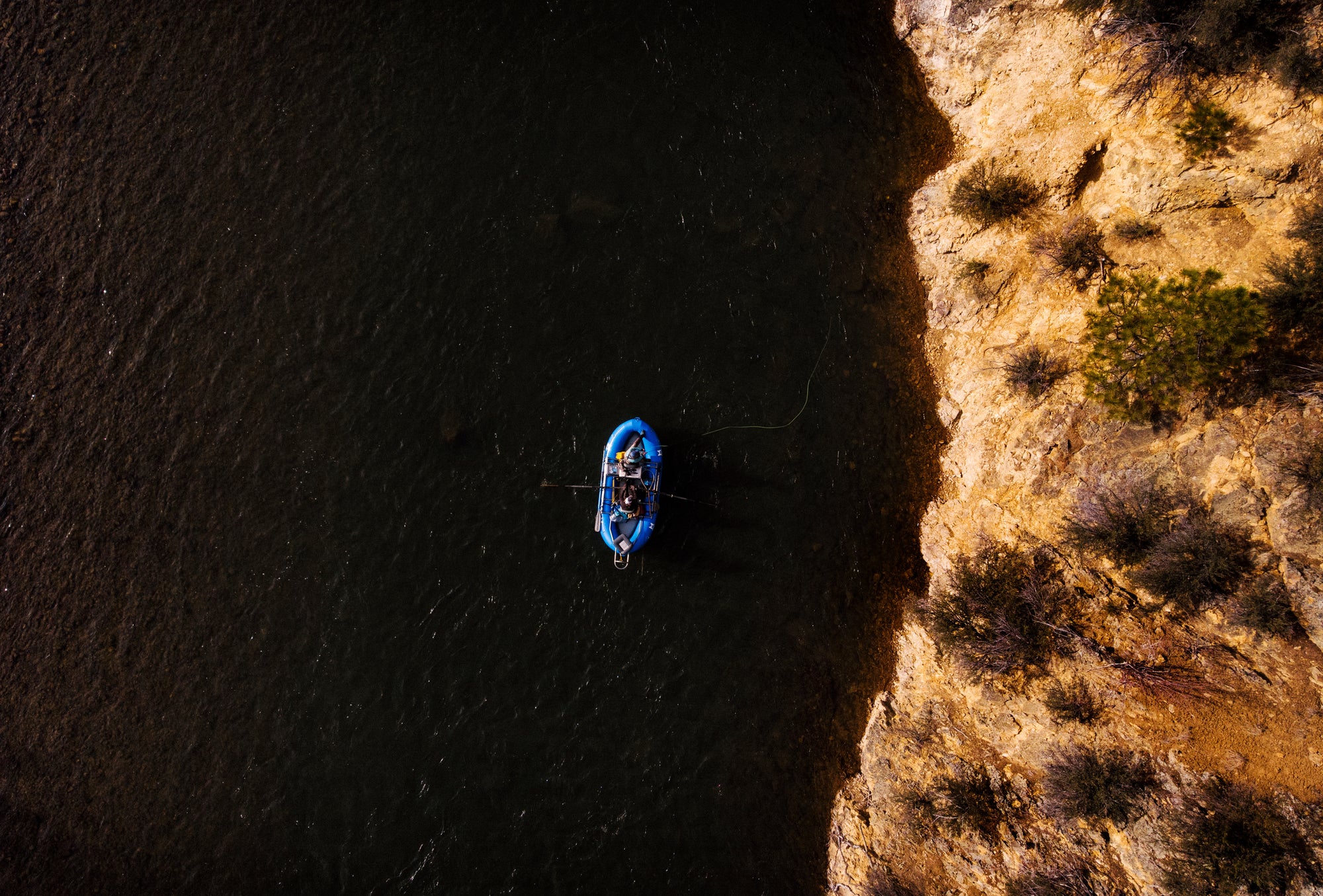 Aerial view of a lone angler fishing from a blue raft near a rocky riverbank with sparse vegetation