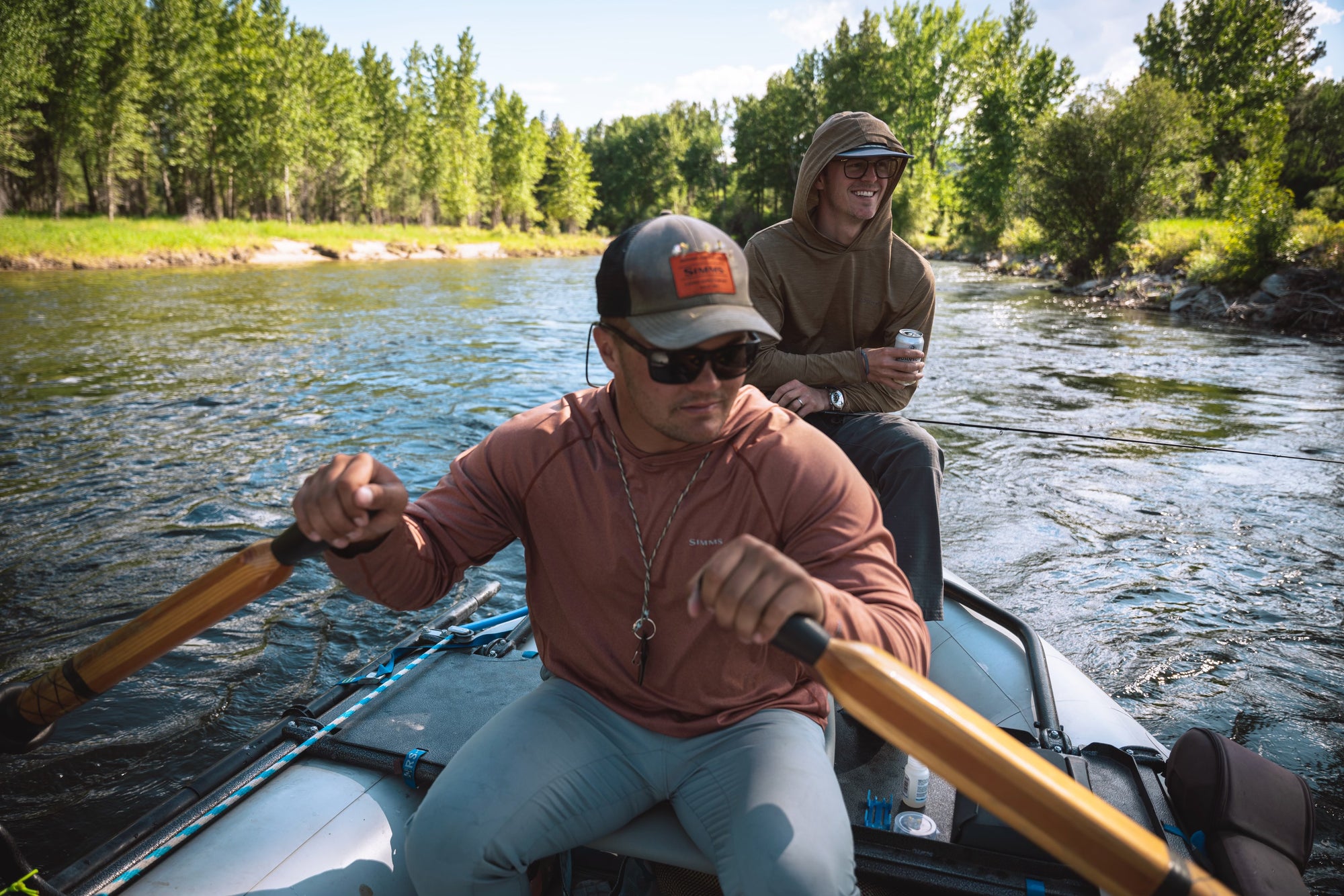 Two anglers in casual outdoor clothing paddling a raft on a river surrounded by green trees under clear sky